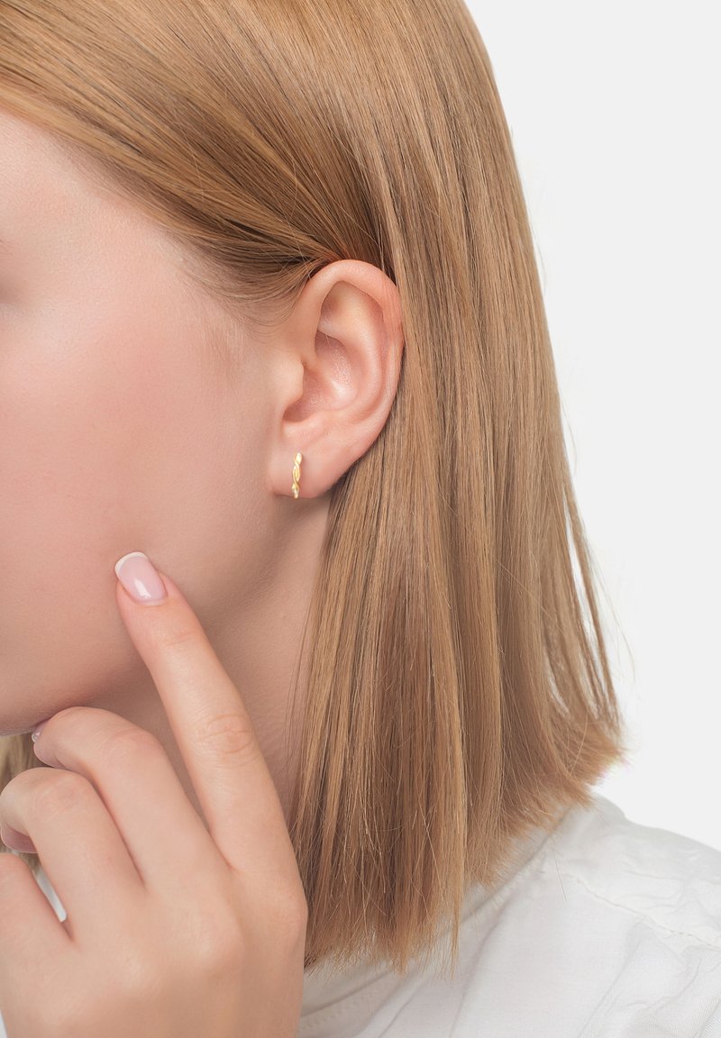 Primo piano dell'orecchio sinistro di una donna con capelli biondi corti, che indossa un piccolo orecchino ad anello attorcigliato in oro, mentre tocca la sua guancia con un dito curato.