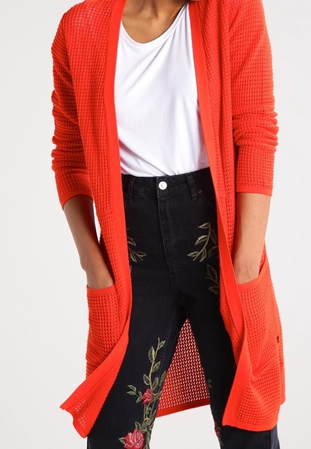 Cardigan rouge en maille ajourée avec manches longues et poches latérales, porté sur un t-shirt blanc. Associé à un pantalon noir avec broderies florales.