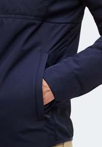 Personne portant une veste bleu marine avec une main glissée dans une poche latérale, mettant en valeur le tissu et les détails des coutures de la veste.