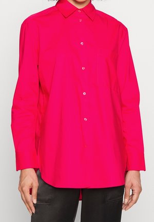 Femme portant une chemise rose vif à manches longues boutonnée avec un col et un pantalon noir, debout devant un fond uni.