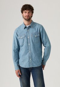 Light blue denim shirt with long sleeves, buttoned front, collar, and two chest pockets. Finished with subtle stitching and a curved hem.