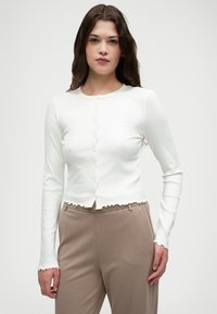 Biały, prążkowany kardigan z długim rękawem, z okrągłym dekoltem, zapięciem na guziki z przodu i falbaniastym wykończeniem, zestawiony z beżowymi spodniami.