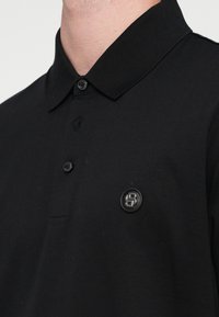 Melnā polo krekls ar teksturētu apkakli, divu pogu aizdari un nelielu apaļu logo kreisajā krūtīs. Gluda kokvilnas auduma.