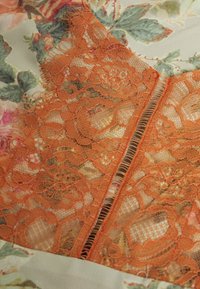 Oranžová krajková tkanina s květinovými vzory položená na krémové tkanině s potiskem zelených listů a růžových květin.