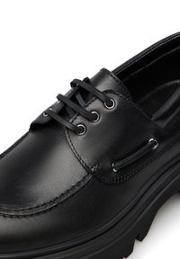 Scarpa in pelle nera con finitura liscia, dotata di lacci passati attraverso occhielli in metallo e suola spessa in gomma. Dettagli di cucitura sottili visibili.