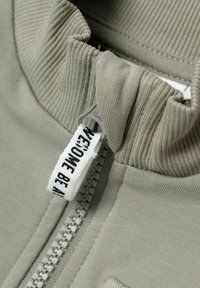 Suéter gris con cremallera y cuello acanalado, con una etiqueta de tela blanca que dice "WE'RE ONE BE" cerca de la cremallera. Textura suave y diseño ajustado.