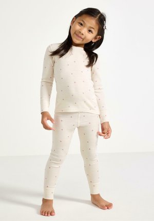 Ung pige med sort hår iført cremefarvet pyjamas med små lyserøde hjerteprint, stående barfodet mod en ensartet lys baggrund.