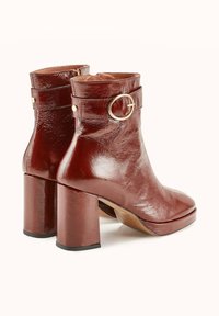 Bottines en cuir brun avec une finition brillante, présentant une boucle dorée ronde en accent sur la sangle et un design de talon épais.