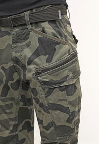 Pantalons cargo camouflage en tons vert et noir, dotés de plusieurs poches, d'un tissu texturé et d'une ceinture contrastante. Détail de fermeture éclair visible.