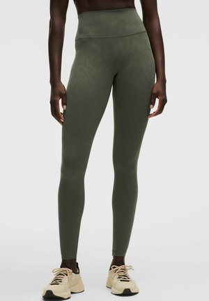 Persona que lleva leggings verdes oliva de talle alto con un patrón sutil y zapatillas deportivas beige, de pie sobre un fondo claro y liso.