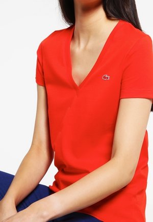 Femme portant un t-shirt rouge à manches courtes avec un col en V et un petit logo crocodile brodé en blanc et rouge sur le côté gauche de la poitrine.