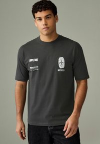 Tricou gri închis cu mânecă scurtă, având text grafic alb și un design circular pe față. Material din bumbac, cu o croială obișnuită.