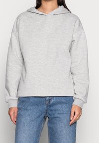 Sudadera gris clara hecha de una suave mezcla de algodón, con una gran capucha, hombros caídos y puños de canalé. Combinada con jeans de mezclilla azules.