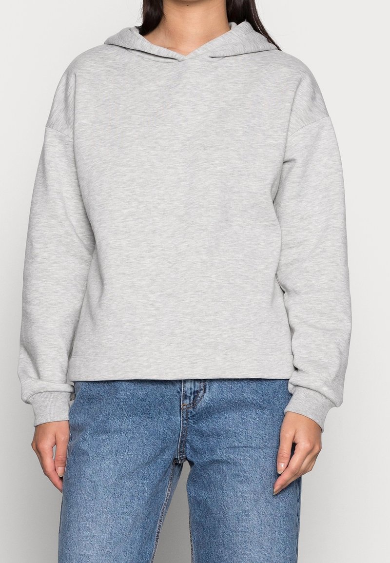 Sudadera gris clara hecha de una suave mezcla de algodón, con una gran capucha, hombros caídos y puños de canalé. Combinada con jeans de mezclilla azules.