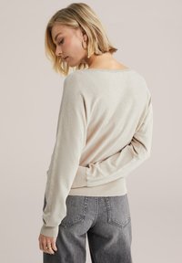 Gai bēšas krāsas pulovers ar brīvu piegriezumu, ribotām piedurknēm un platu apkakli, kombinēts ar tumši pelēkiem augstās jostasvietas biksēm.