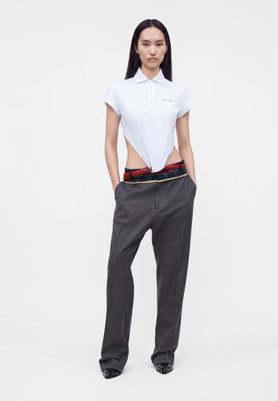 Jeune femme aux longs cheveux foncés porte un polo blanc à manches courtes rentré dans un pantalon gris ample avec une ceinture rouge, debout les mains dans les poches.