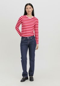 Rood en roze gestreepte langarmshirt gecombineerd met donkerblauwe spijkerbroek. Model draagt zwarte schoenen en poseert tegen een neutrale achtergrond.