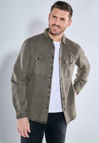 Olivgrüne Knopfhemd mit zwei Brusttaschen und langen Ärmeln, aus weichem Stoff; über einem weißen T-Shirt getragen und mit dunklen Hosen kombiniert.
