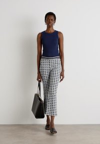 Marineblauwe mouwloze top, gestreepte zwart-witte broek, zwarte tote bag en zwarte slippers. Witte achtergrond, neutrale vloer.