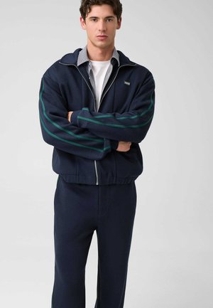 Junger Mann steht mit verschränkten Armen, trägt einen marineblauen Trainingsanzug mit grünen Streifen und eine teilweise zugezogene Jacke über einem weißen Hemd.