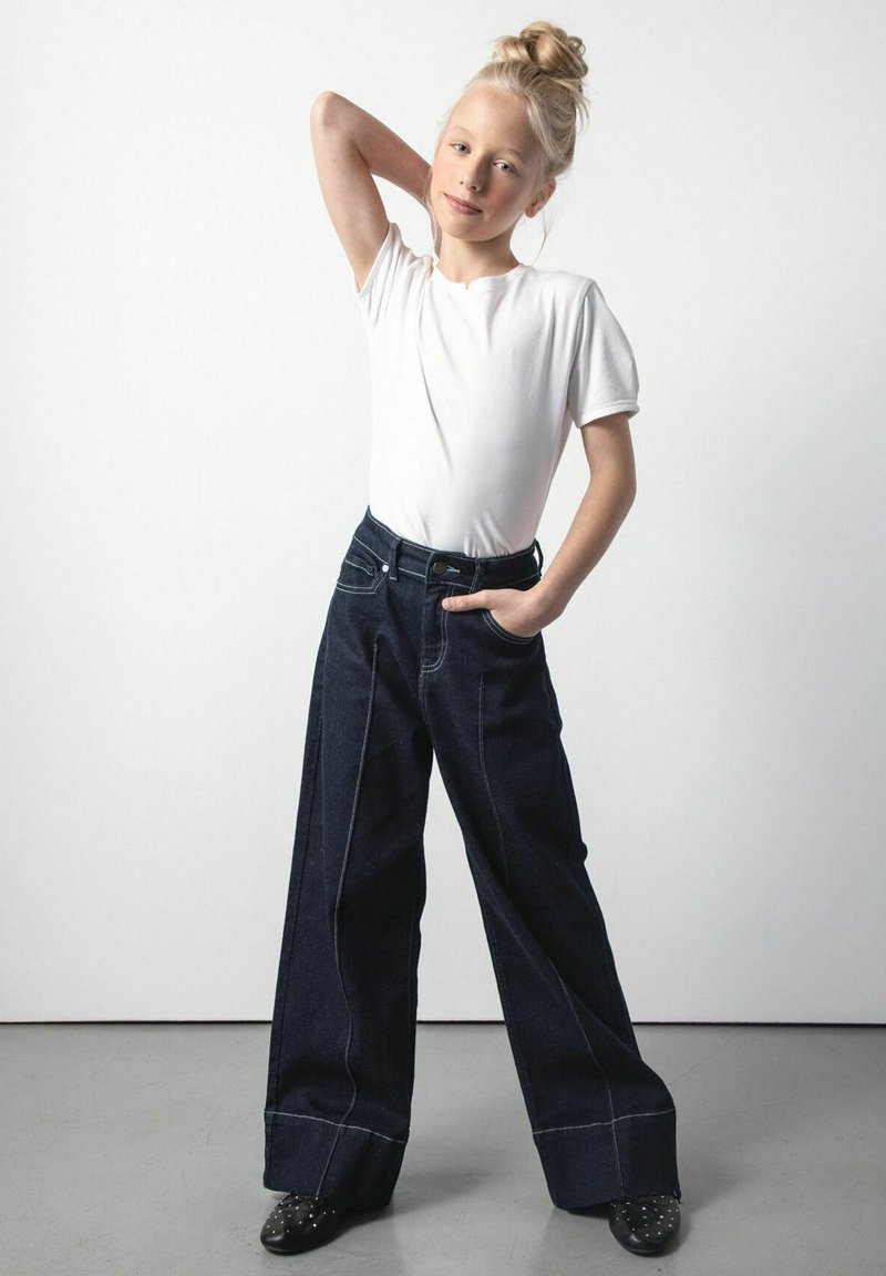 Biała koszulka z krótkim rękawem w połączeniu z ciemnoniebieskimi spodniami o szerokich nogawkach z przednimi kieszeniami i kontrastowym przeszyciem. Całość uzupełniają czarne buty z ćwiekami.