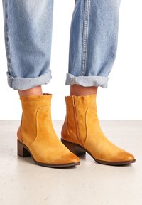 Semišové kotníkové boty v hořčicově žluté barvě s bočním zipem, špičatou špičkou a nízkým stohovaným podpatkem. Rolované světlé modré džíny nad botami.