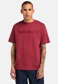 Camiseta de algodón color marrón con el logotipo bordado de "Timberland" y un icono de árbol en el frente, con mangas cortas y un escote redondo.