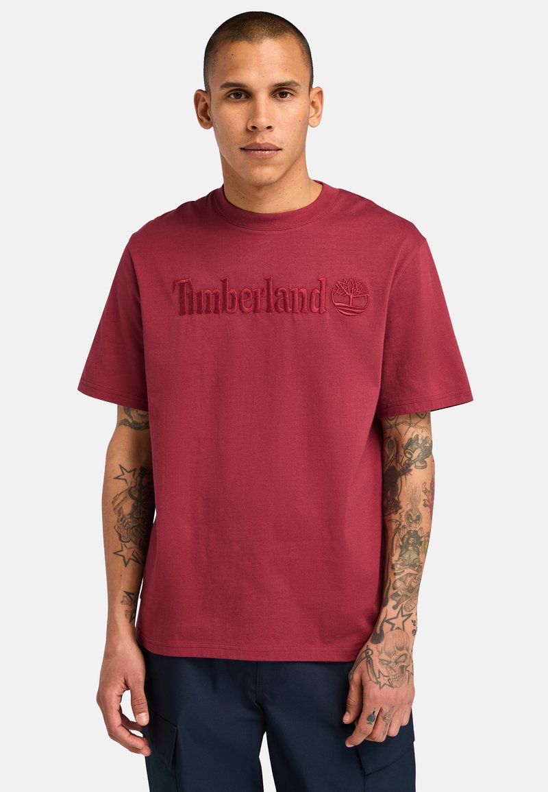 Camiseta de algodón color marrón con el logotipo bordado de "Timberland" y un icono de árbol en el frente, con mangas cortas y un escote redondo.