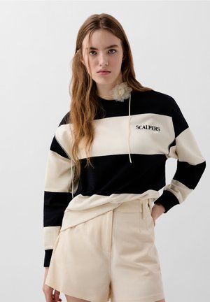 Felpa a righe nera e crema con cappuccio e logo; abbinata a pantaloni corti beige. La felpa presenta una texture morbida e una vestibilità rilassata.