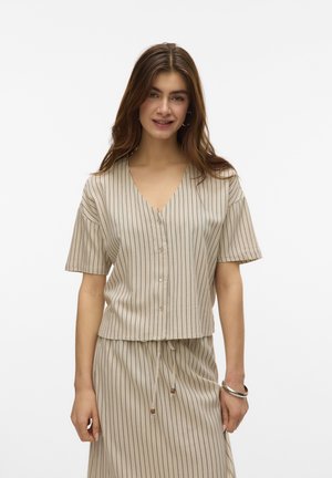 Joven mujer con blusa beige de rayas finas de manga corta y botones, y falda a juego con cordón, de pie frente a fondo blanco.
