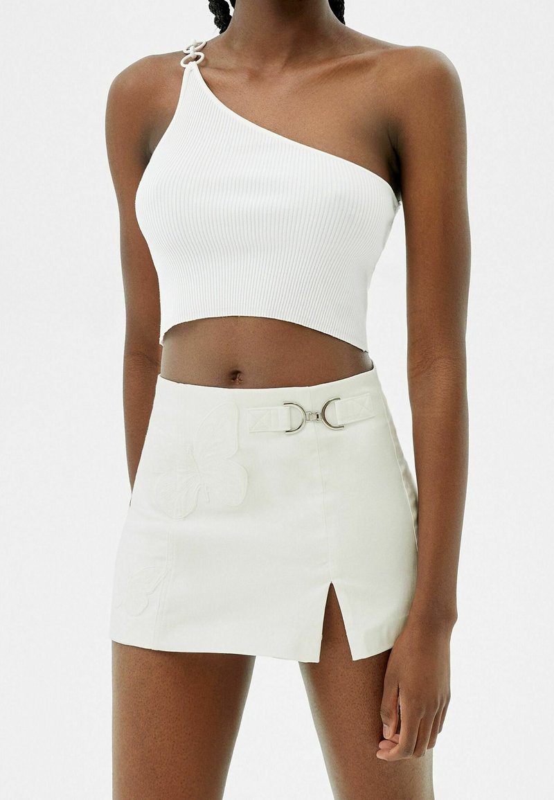 Femme portant un crop top blanc à une épaule et une mini jupe blanche avec une fente devant et un détail boucle métallique.