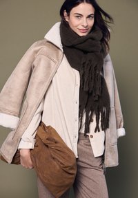 Giacca in shearling beige con fodera bianca, sciarpa frangiata marrone scuro, camicia in corduroy crema, borsa in pelle marrone e pantaloni marrone a fantasia.