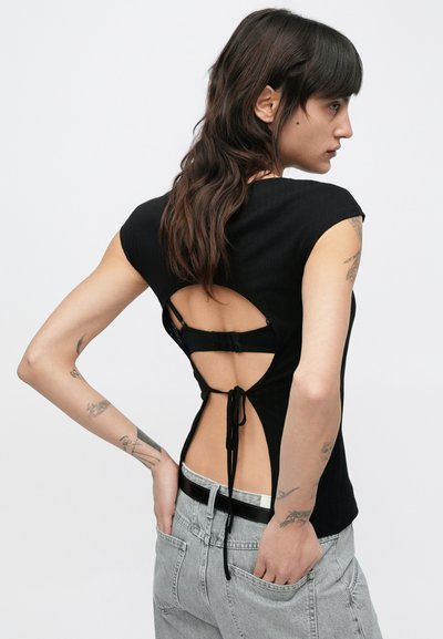 Haut noir côtelé à manches courtes, avec un dos découpé, une fermeture à nouer réglable, et un tissu texturé. Porté avec un jean gris clair.