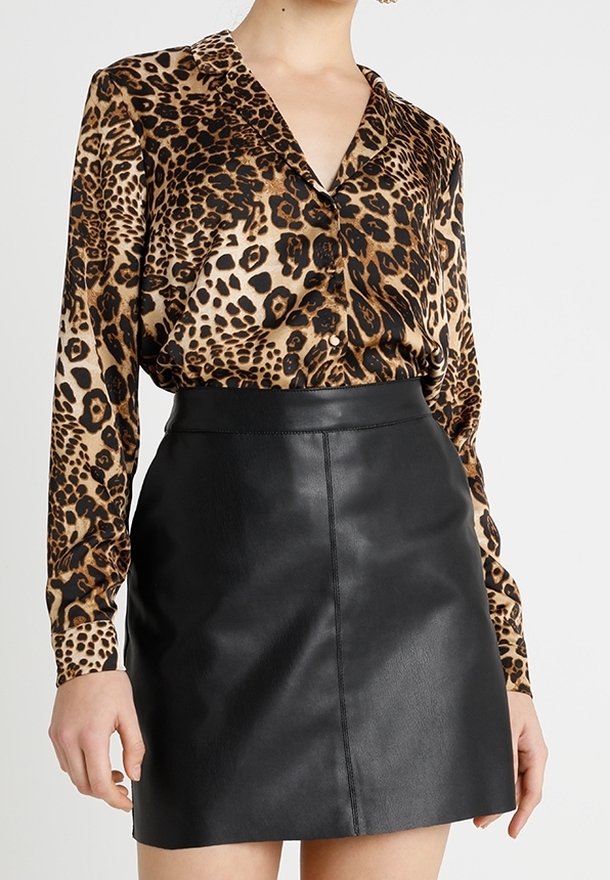 Femme portant un chemisier à manches longues à imprimé léopard, rentré dans une mini-jupe en cuir noir taille haute, sur un fond uni.