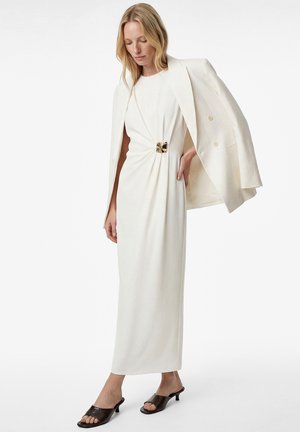 Kobieta w długiej białej sukni z ozdobą w postaci złotej klamry, białą marynarką zarzuconą na ramiona oraz czarnymi sandałami na obcasie z odkrytymi palcami.