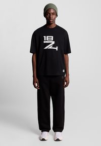 Schwarzes T-Shirt mit großem weißem Grafikdruck, kombiniert mit locker sitzenden schwarzen Hosen. Das Modell trägt eine grüne gestrickte Mütze und hellfarbene Sneakers.