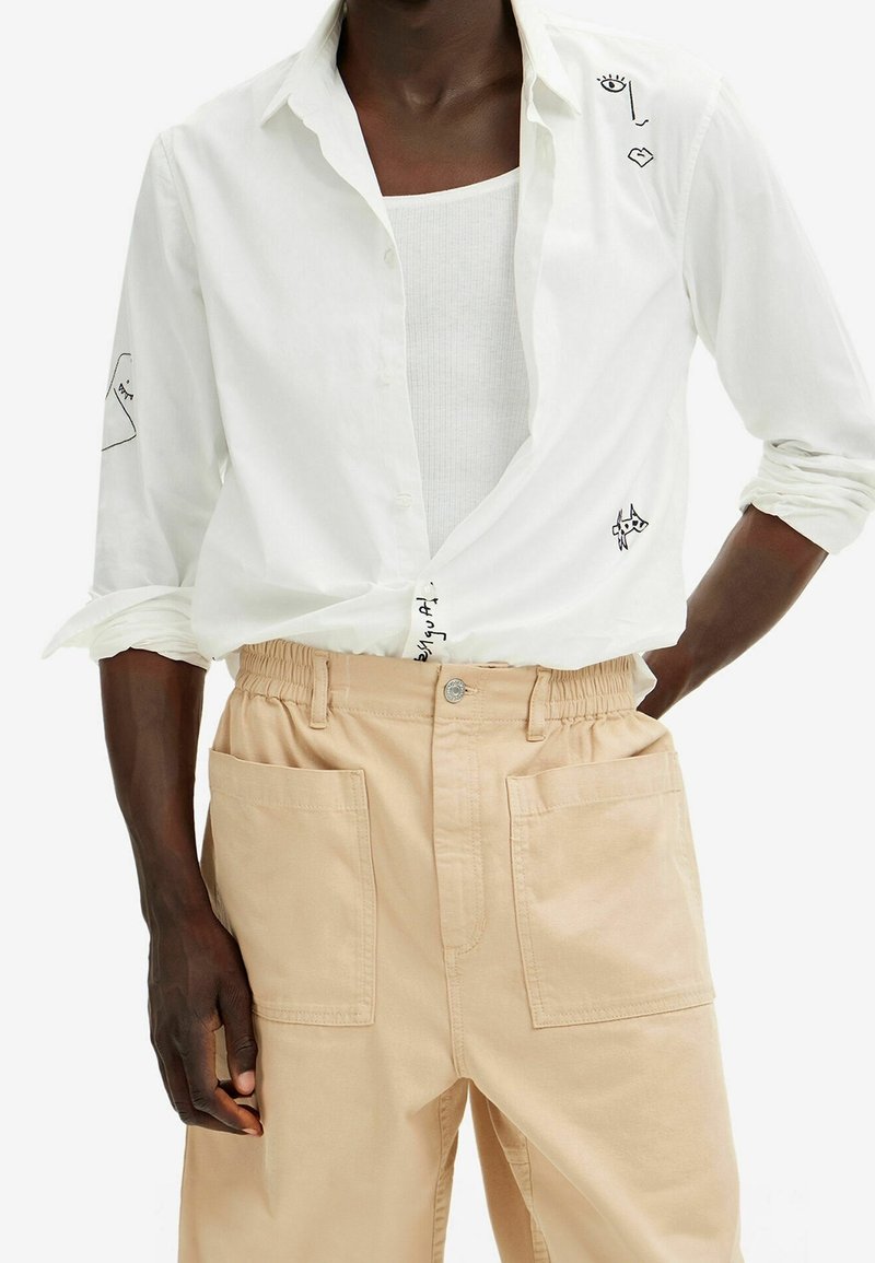 Camisa blanca de botones con diseños abstractos en negro, llevada sobre una camiseta sin mangas gris claro. Combinada con pantalones cargo beige de cintura elástica y con bolsillos.