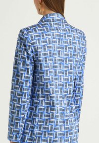 Veste à motifs bleus présentant un design géométrique avec des formes bleu clair et bleu foncé alternées ; texture lisse et coupe structurée.