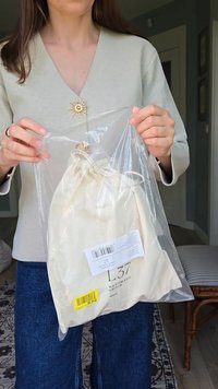Krämfärgad dragsko-väska i transparent plastförpackning, med en varumärkesetikett och ett guldaccent vid stängningen.