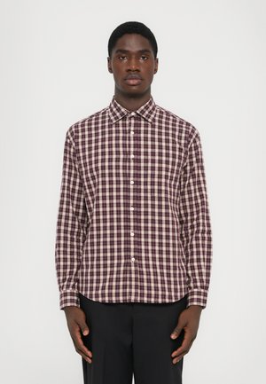 Hombre de pie contra un fondo blanco, vistiendo una camisa de cuadros en tonos burdeos y blancos con pantalones negros.