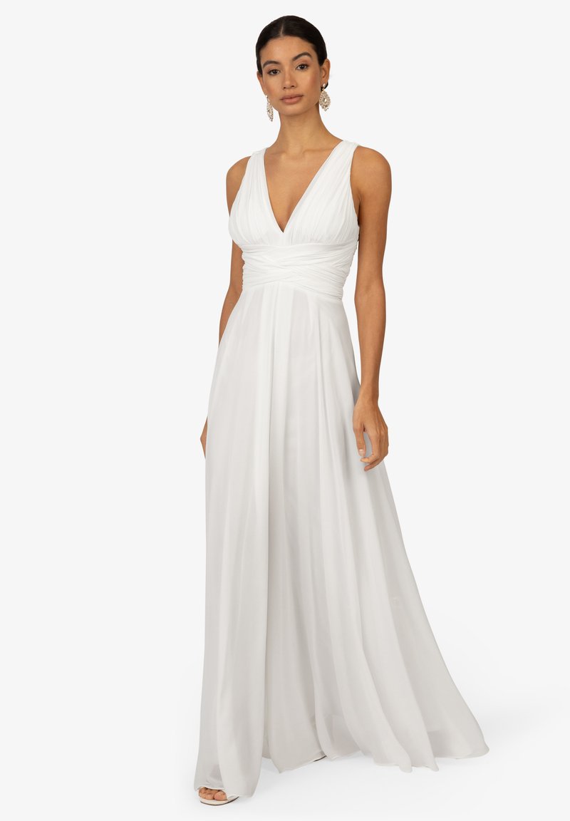 Robe blanche avec un décolleté en V profond, un corsage plissé et une jupe fluide. Dispose de larges bretelles et d'une texture de tissu lisse et légère.
