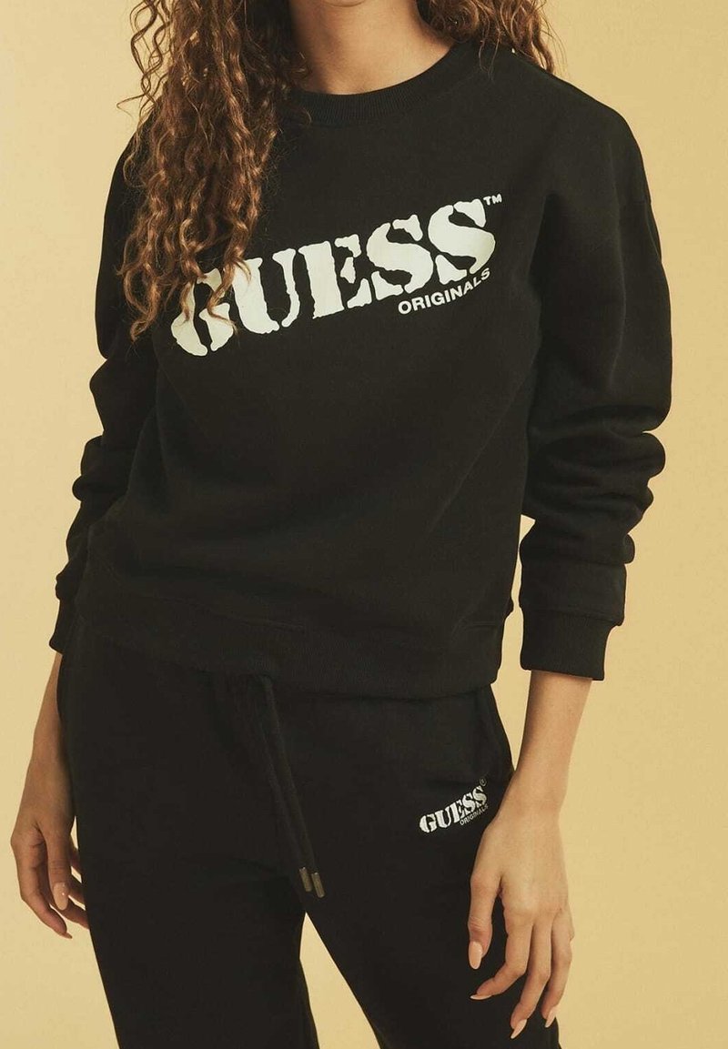 Zwart sweatshirt met wit "GUESS ORIGINALS" logo, geribbelde manchetten en hals. Gecombineerd met bijpassende zwarte broek met een klein logo.