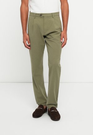 Mann trägt olivgrüne plissierte Hose, braune Loafers und ein weißes, eingestecktes Hemd, steht vor einem schlichten weißen Hintergrund.