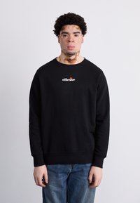 Sudadera negra con cuello redondo y puños acanalados. Presenta un logo blanco de "ellesse" en el pecho. Se lleva sobre un vaquero de mezclilla azul.