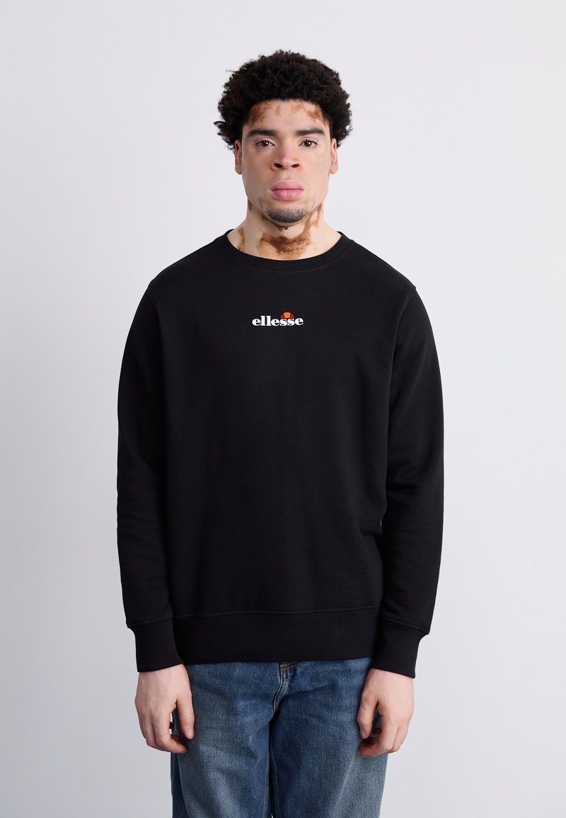 Sudadera negra con cuello redondo y puños acanalados. Presenta un logo blanco de "ellesse" en el pecho. Se lleva sobre un vaquero de mezclilla azul.