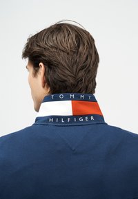 Άνδρας με καστανά μαλλιά που φοράει μπλε σκούρο πουκάμισο με όρθιο γιακά, διακοσμημένο με μπλοκ χρωμάτων κόκκινου, λευκού και μπλε και κείμενο "Tommy Hilfiger".