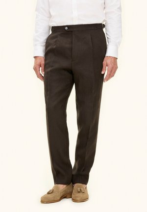 Uomo che indossa pantaloni sartoriali marrone scuro, camicia bianca elegante e mocassini chiari con nappine, in piedi davanti a uno sfondo neutro.