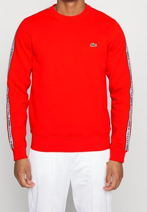 Rød bomuldssweatshirt med rund hals, lange ærmer og kontrasterende sorte og hvide mønstrede sidelister. Har en lille grøn logo på brystet.