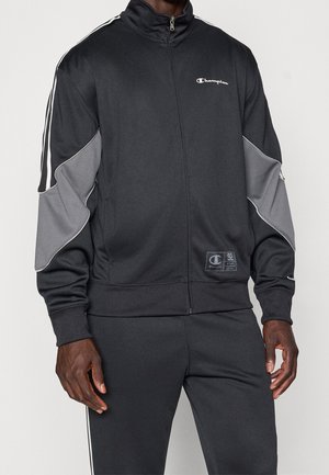 Homme portant une veste de survêtement noire et grise Champion et un pantalon noir assorti avec des bandes blanches sur les manches et les côtés.