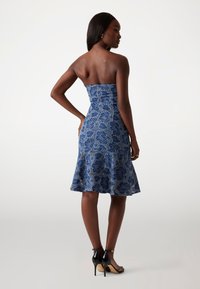 Robe bleue sans bretelles avec des motifs floraux, corsage ajusté, ourlet évasé et texture semblable à de la dentelle. Portée avec des sandales à talons hauts noires.